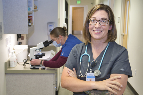 Medical assistant (MA) Tambria Cox stands with arms folded, smiling, while colleague works in the background