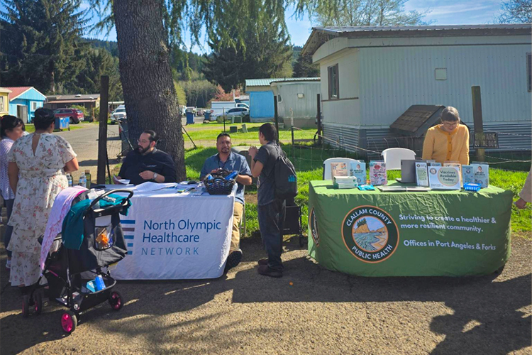 Tables representing local health care organizations, next to each other under a tree, with staff and public in view.