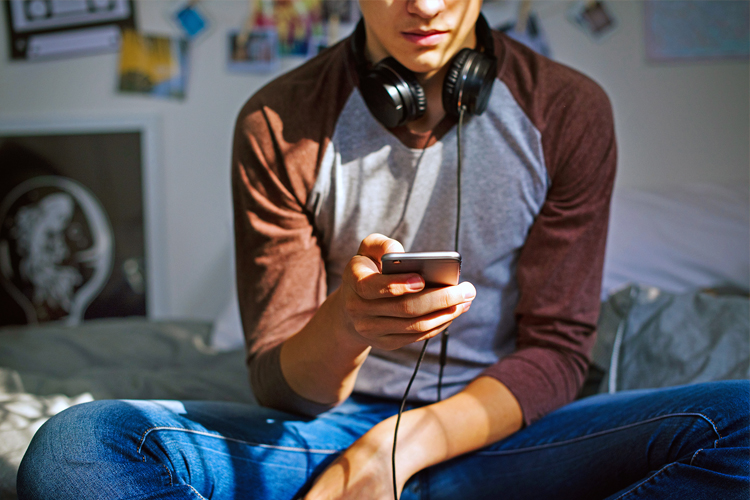 Teen with headphones around his neck, sitting on bed using smartphone